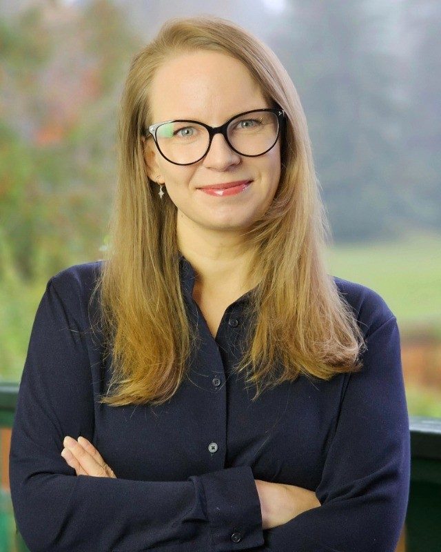 Olga Mazur, smiling, with glasses and a navy shirt, standing outdoors with her arms crossed
