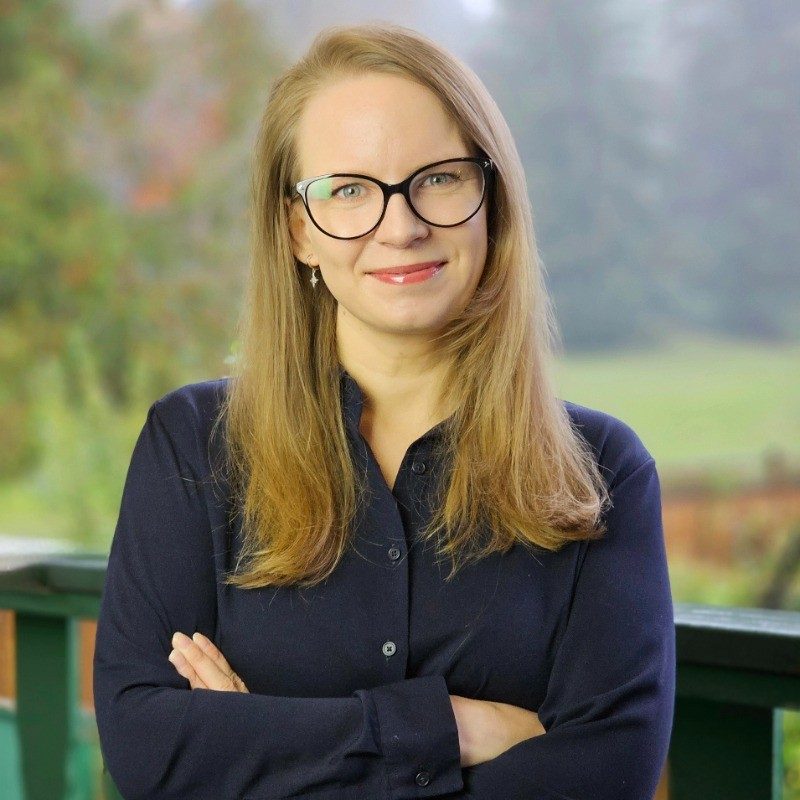 Olga Mazur, smiling, with glasses and a navy shirt, standing outdoors with her arms crossed